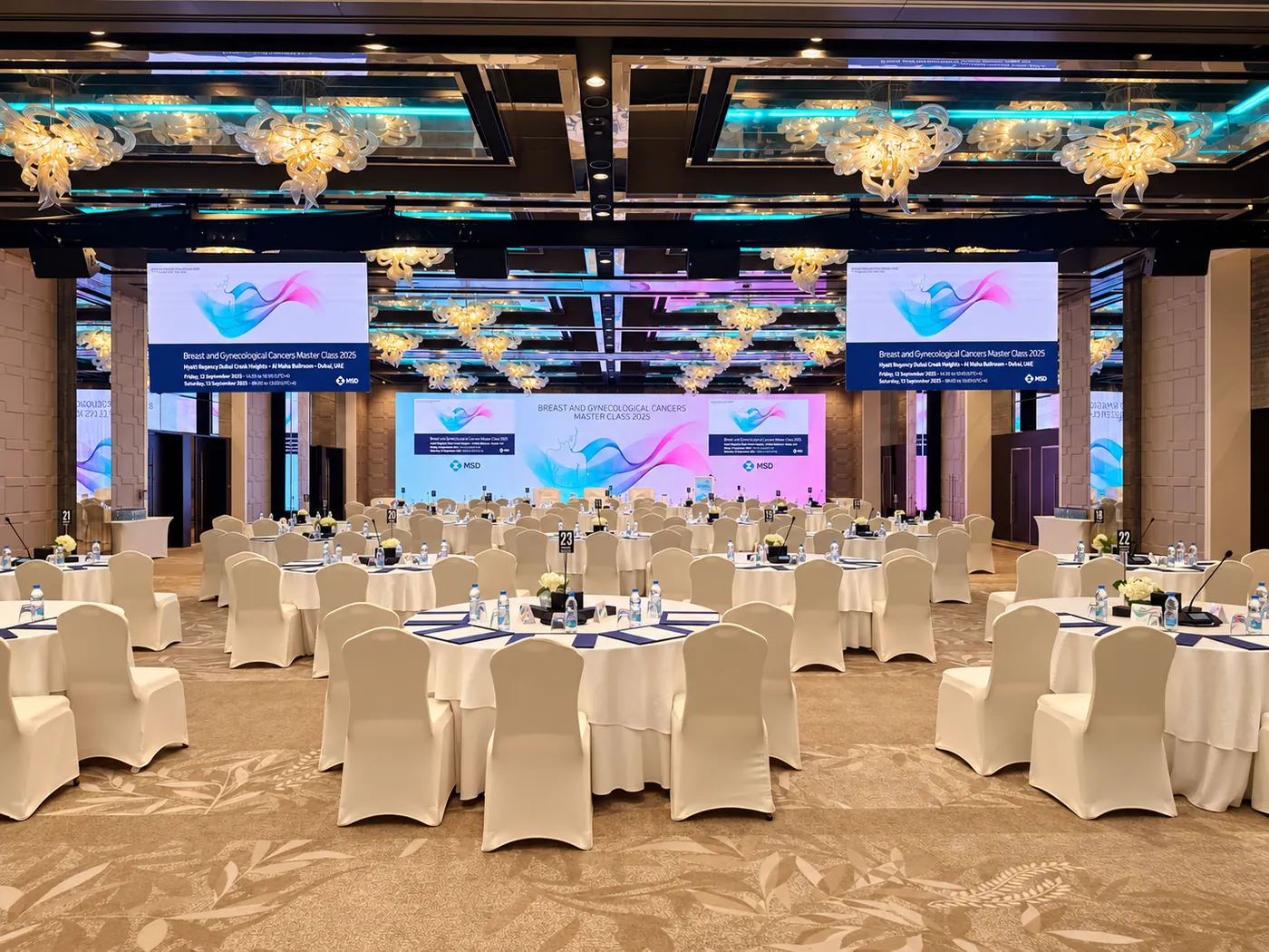 A medical conference setup with a wide presentation backdrop, suspended screens and round-table seating.
